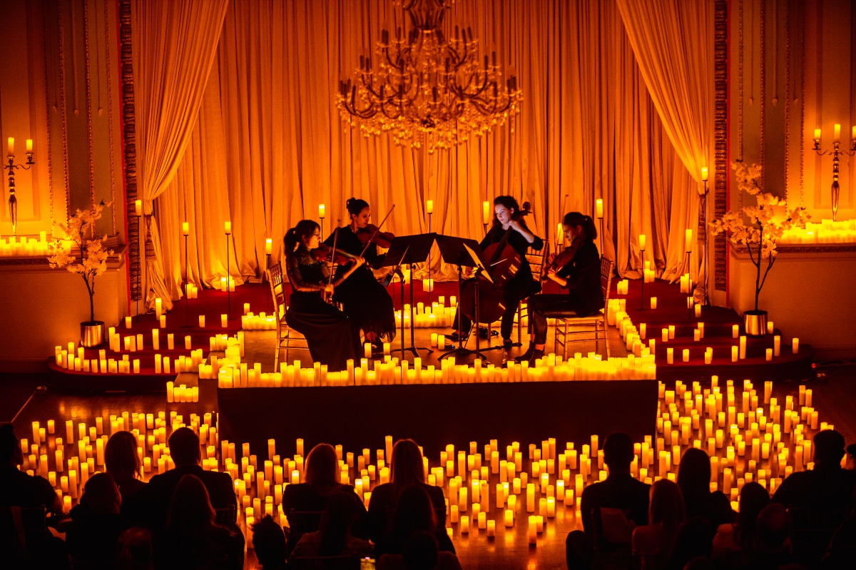 A string quartet performing on a stage surrounded by candles and the silhouette of an audience in front. 
